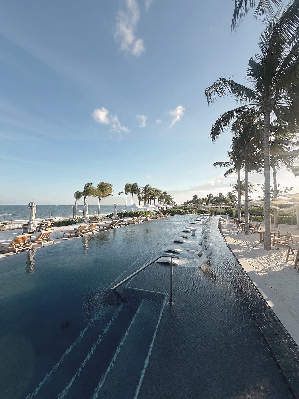 Black-tile pool with in-water loungers, palms and the sea at the far edge