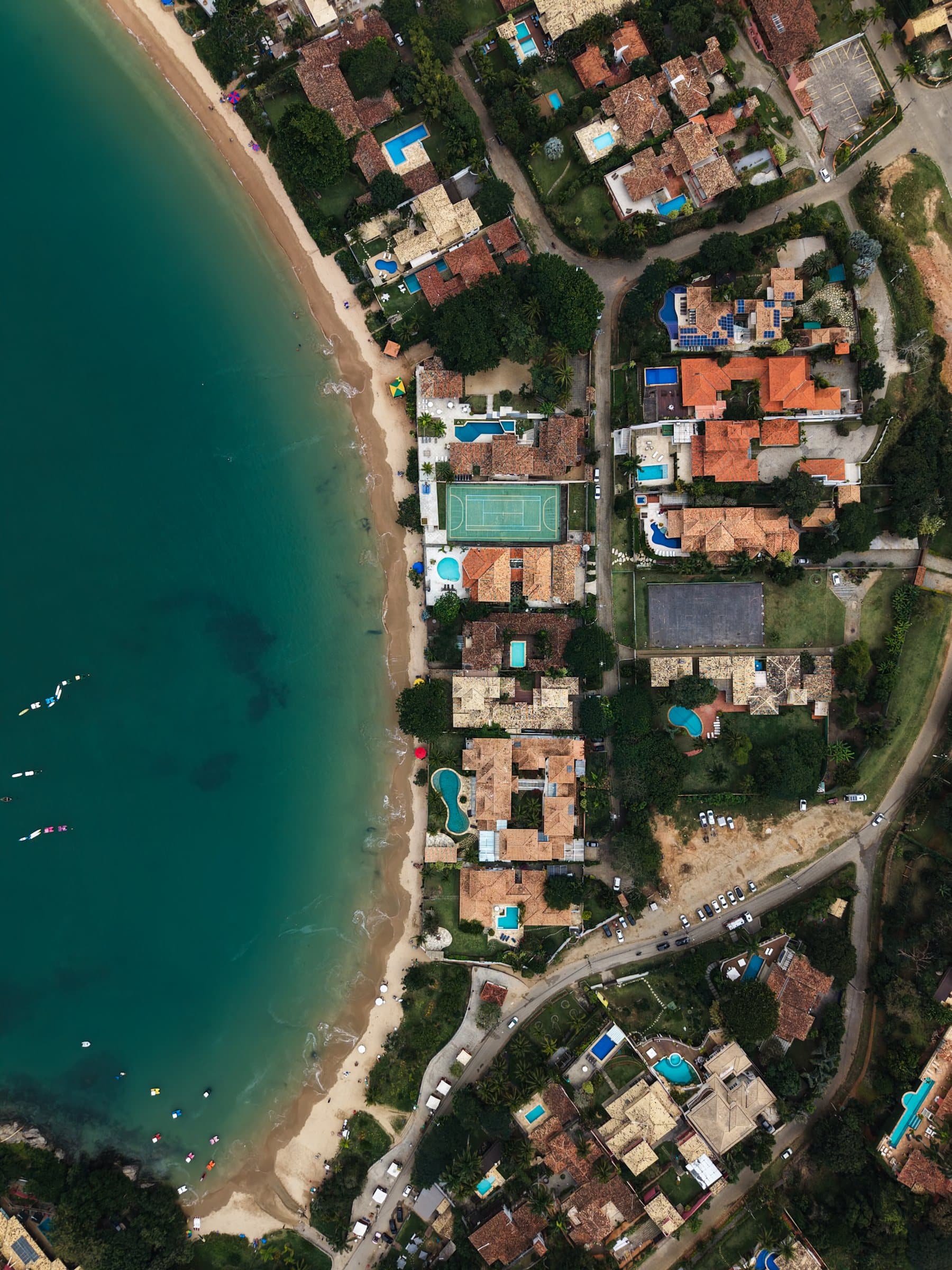 Top-down view of hillside houses and pools beside a curving beach